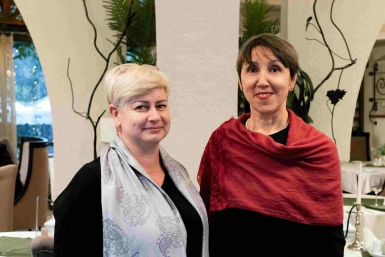 Aliona stands on the left of the image wearing a grey scarf, with her blonde hair parted to the side. On the right of the image Aliona's friend Maria stands wearing a red scarf - both ladies are wearing black tops and smiling into the camera.